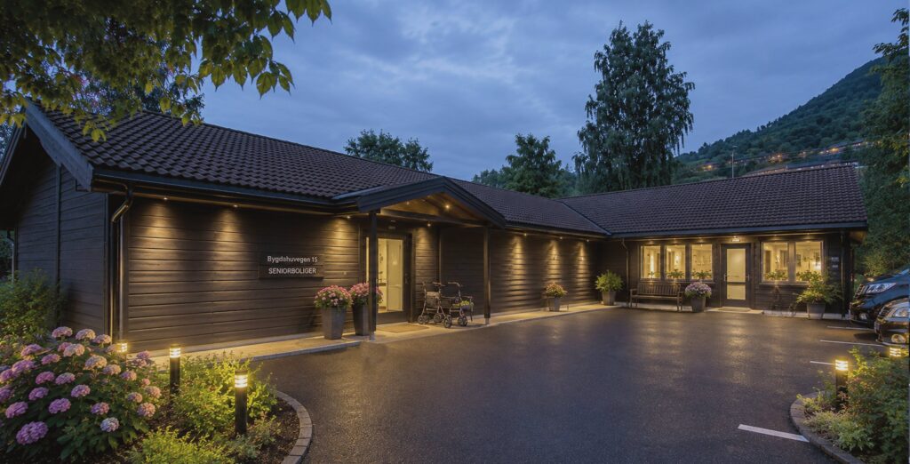 Visualisering av det ferdige bygget i skumring, sett fra inngangssiden, med opplyste vinduer, blomsterkasser og skiltet «Bygdahusvegen 15 – seniorboliger».
